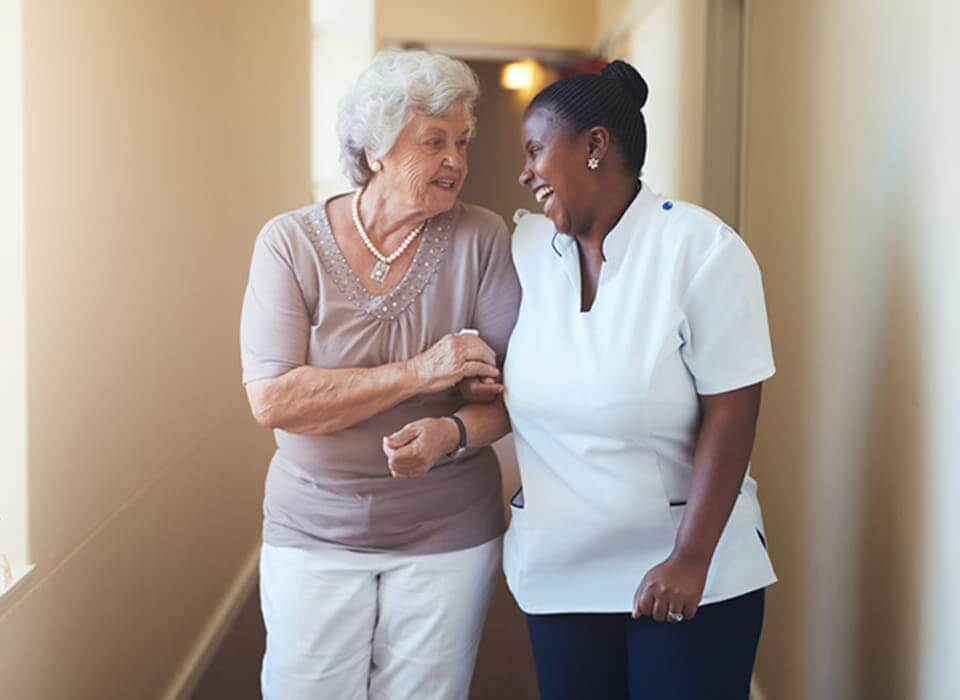 Caregiver laughs and smiles with elderly female client as they walk in client’s home.