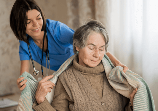Young nurse in blue scrubs helps elderly woman get comfortable by draping a blanket over her.