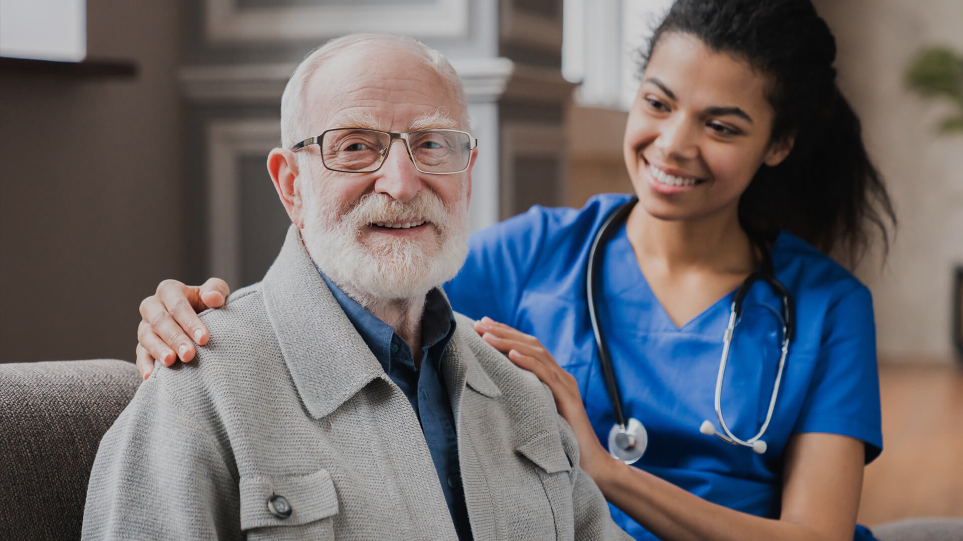 Smiling nurse in blue scrubs comforts a smiling elderly male patient.