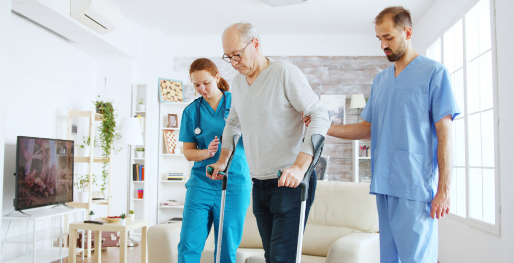 Two home health nurses helping an older man walk with forearm crutches in a bright living room
